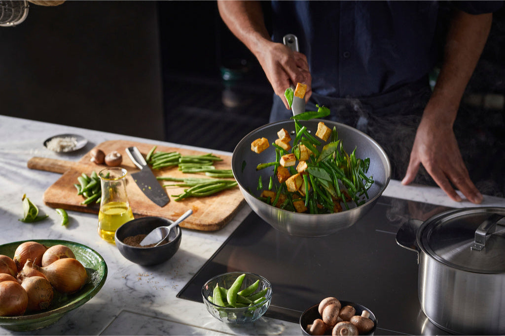 Person lagar grönsakswok i panna från Nordic Cookware på marmorbänk i modernt kök